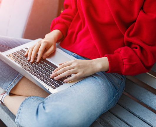 Crop young female in casual red blouse and jeans sitting on bench and typing on laptop while browsing internet and searching information